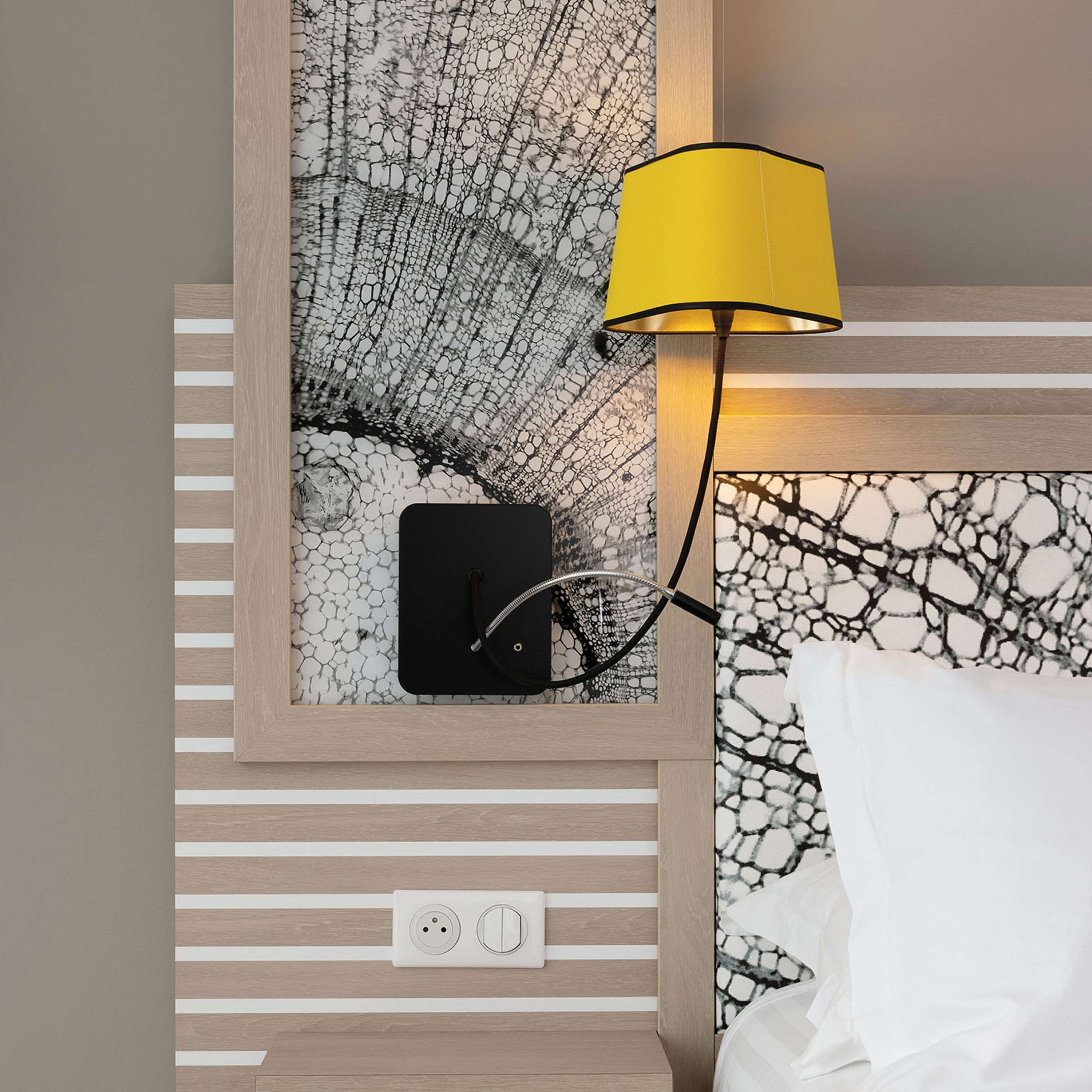 A hotel room with a bed, a nightstand with a lamp, a framed picture, and a power outlet.