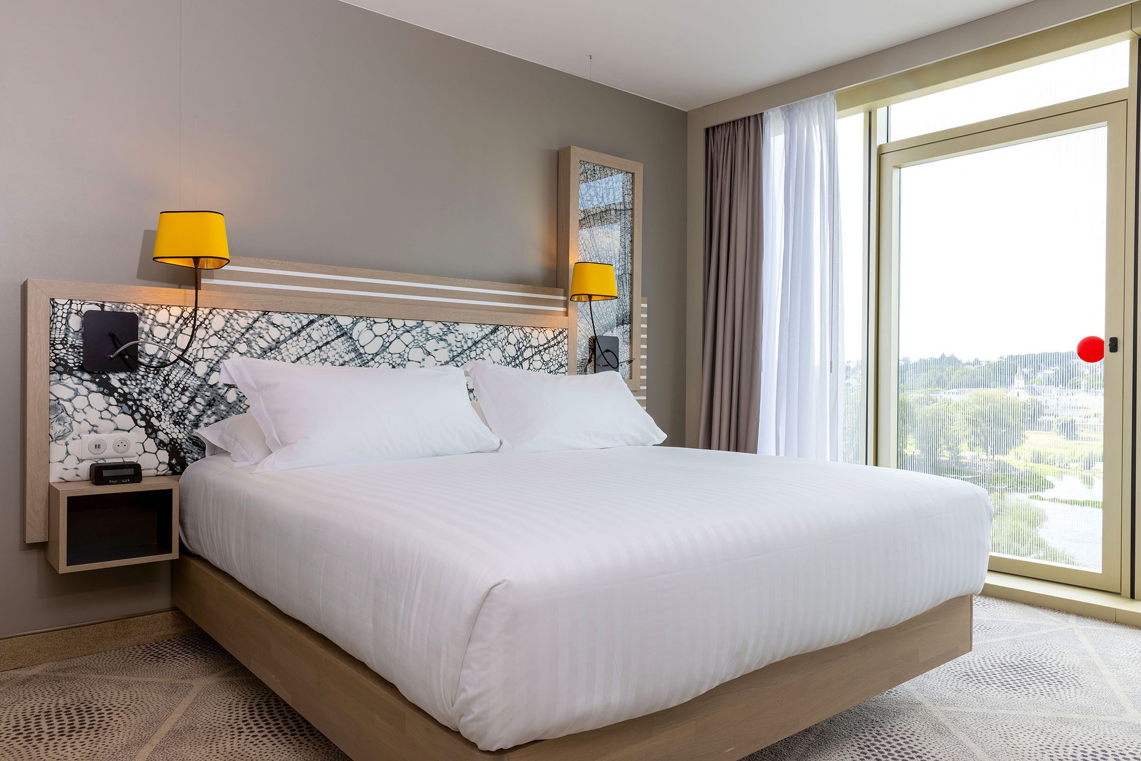 A hotel room with a large bed, two bedside lamps, a nightstand with a clock, and a view of the city through a large window.