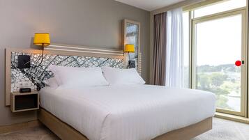 A hotel room with a large bed, two bedside lamps, a nightstand with a clock, and a view of the city through a large window.
