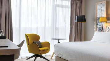 Habitación de hotel con cama, escritorio, silla, lámpara y vistas a través de cortinas traslúcidas.