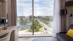 A modern hotel room with a large window, a sofa, a desk with a lamp, and a view of a bridge and cityscape. - Hilton Garden Inn Tours Centre (Tours)