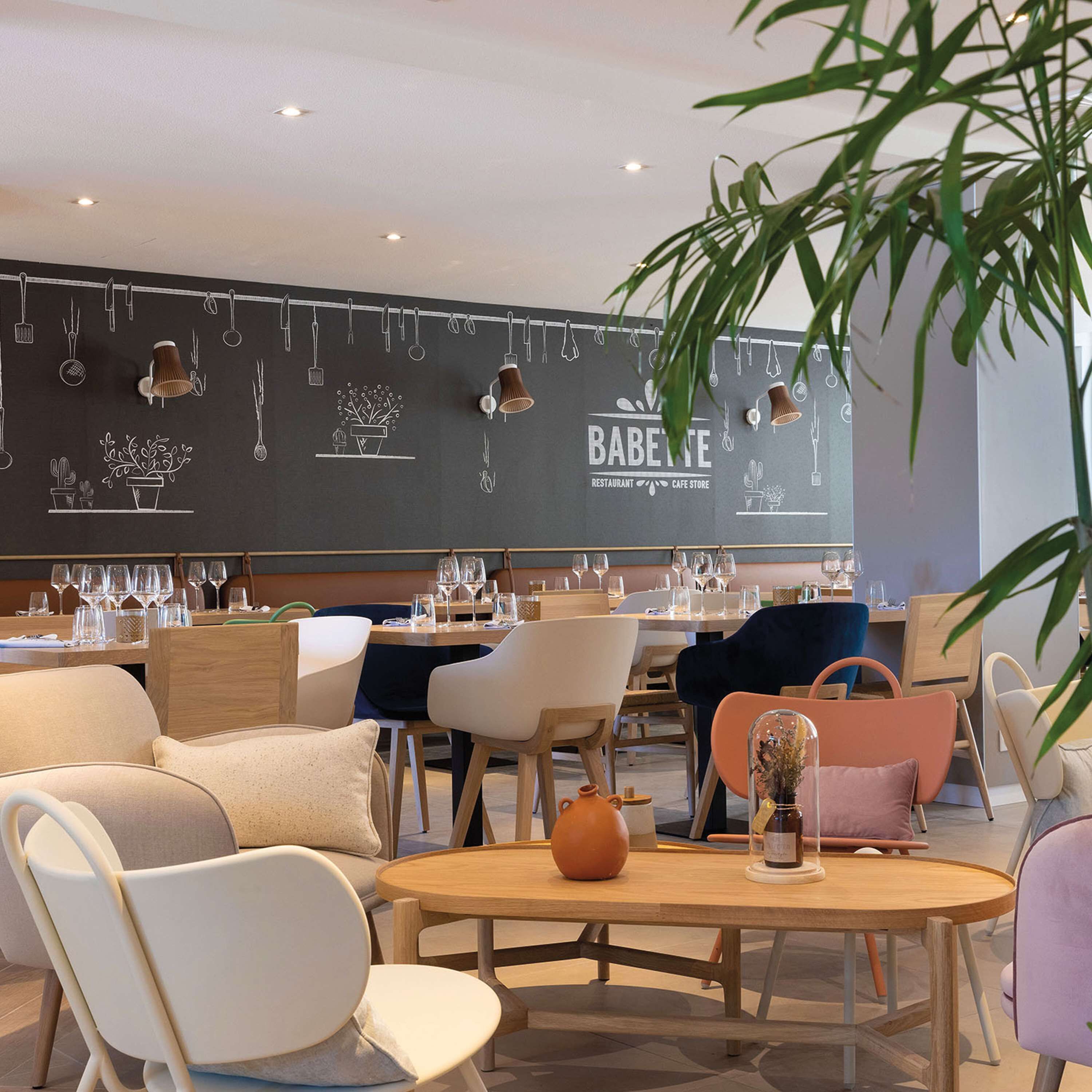 Interior moderno de restaurante con una pared de pizarra mostrando utensilios y plantas, mesas y sillas de madera.