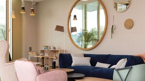A modern living room with a blue sofa, a pink chair, and a teal chair. There are two round mirrors on the wall, a floor lamp, and a bookshelf with various items. - Hilton Garden Inn Tours Centre (Tours)