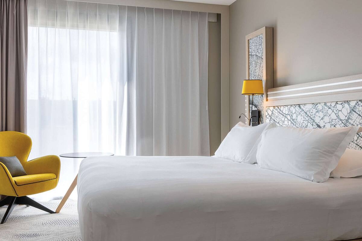A hotel room with a large bed, a yellow armchair, a side table, and a window with sheer curtains.