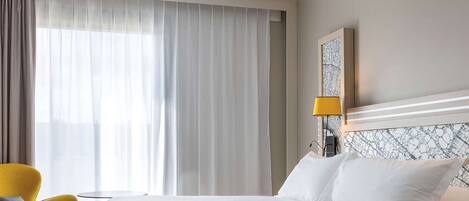 Een hotelkamer met een groot bed, een gele fauteuil, een bijzettafeltje en een raam met dunne gordijnen.