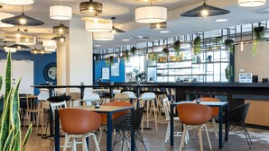 A modern restaurant interior with multiple dining tables and chairs, hanging plants, and a bar area with shelves of bottles. - Hampton by Hilton Tours Centre (Tours)