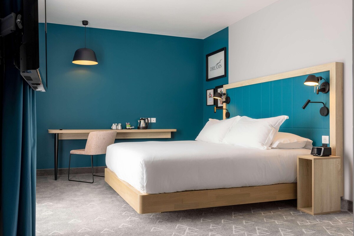 Una habitación de hotel moderna con una pared de acento color esmeralda, un cabecero de madera, un escritorio con silla y una mesita de noche con lámpara.