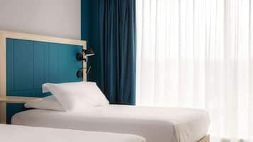 Habitación de hotel con dos camas individuales, cabecera de madera, mesita de noche, lámpara de escritorio y un ventanal con cortinas traslúcidas.