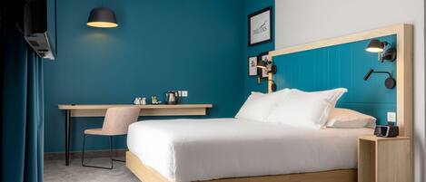 A modern hotel room with a teal accent wall, a wooden bed frame, a desk with a chair, and a nightstand with a lamp.