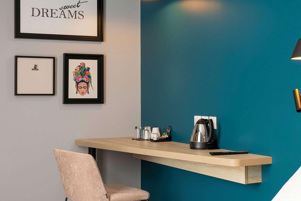 Una habitación moderna con paredes color esmeralda, un escritorio de madera, una silla y una cama. Hay cuadros enmarcados en la pared, una tetera en el escritorio y una lámpara de techo.