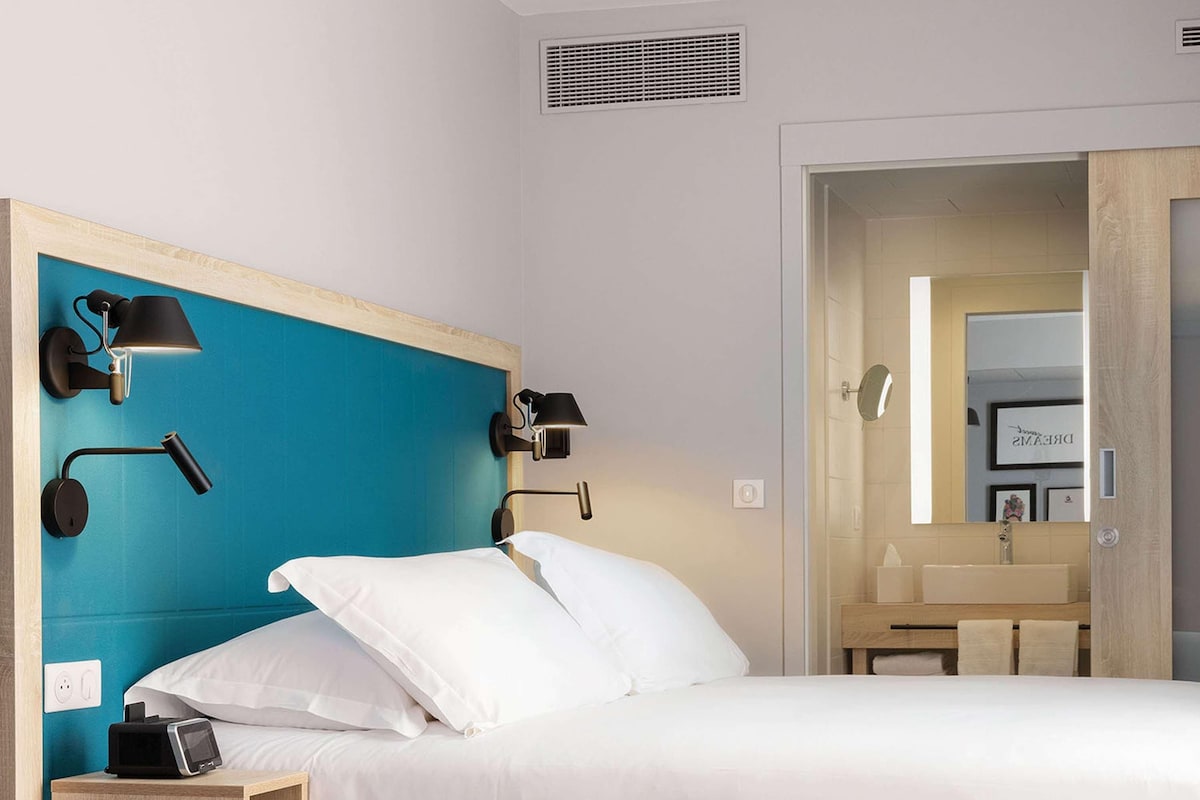 Una habitación de hotel moderna con una cama, una mesita de noche, un escritorio y un baño visible a través de una puerta abierta.