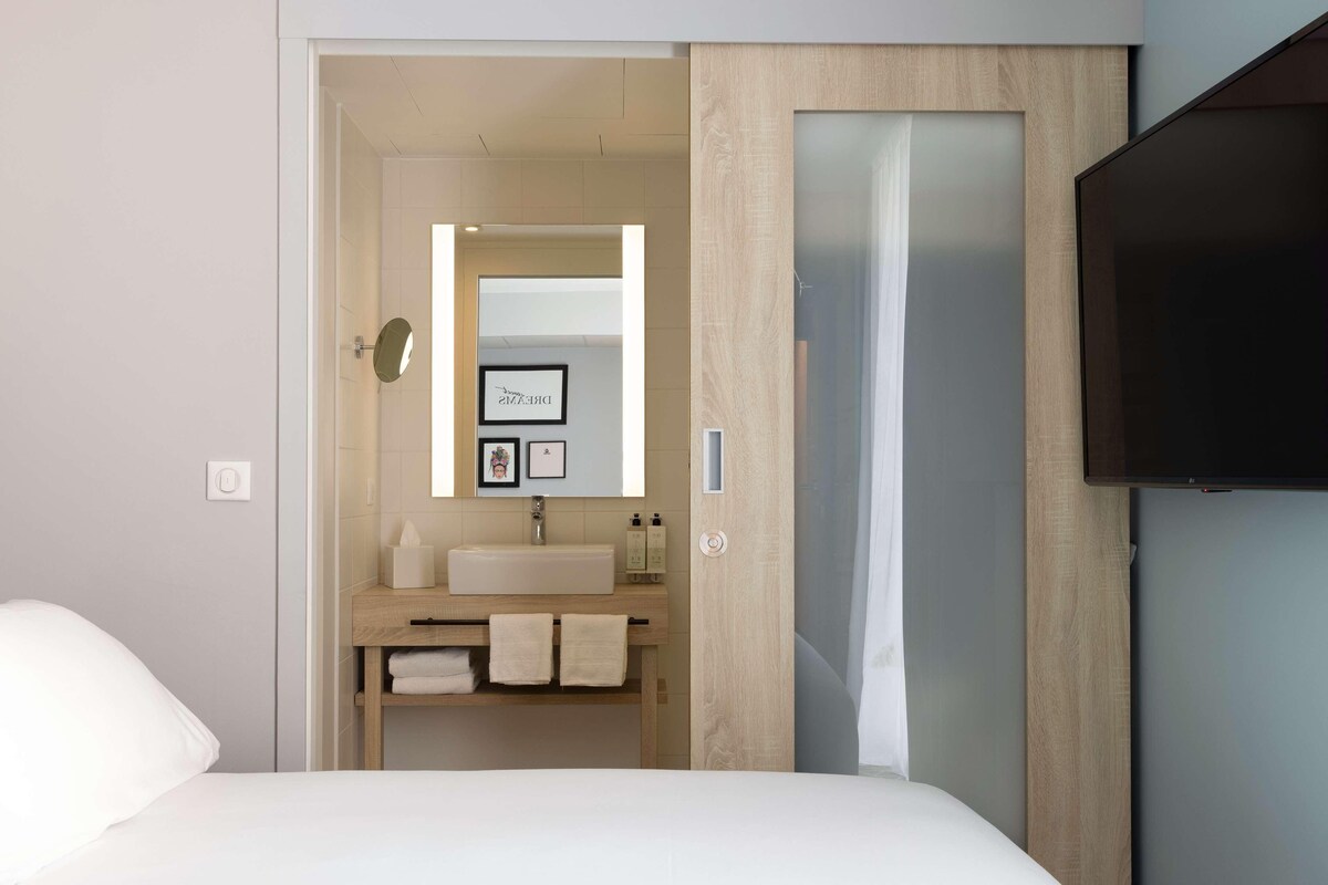 Una habitación de hotel moderna con una cama grande, un armario de madera, un televisor de pantalla plana, un baño con lavamanos y espejo, y un cuadro enmarcado en la pared.
