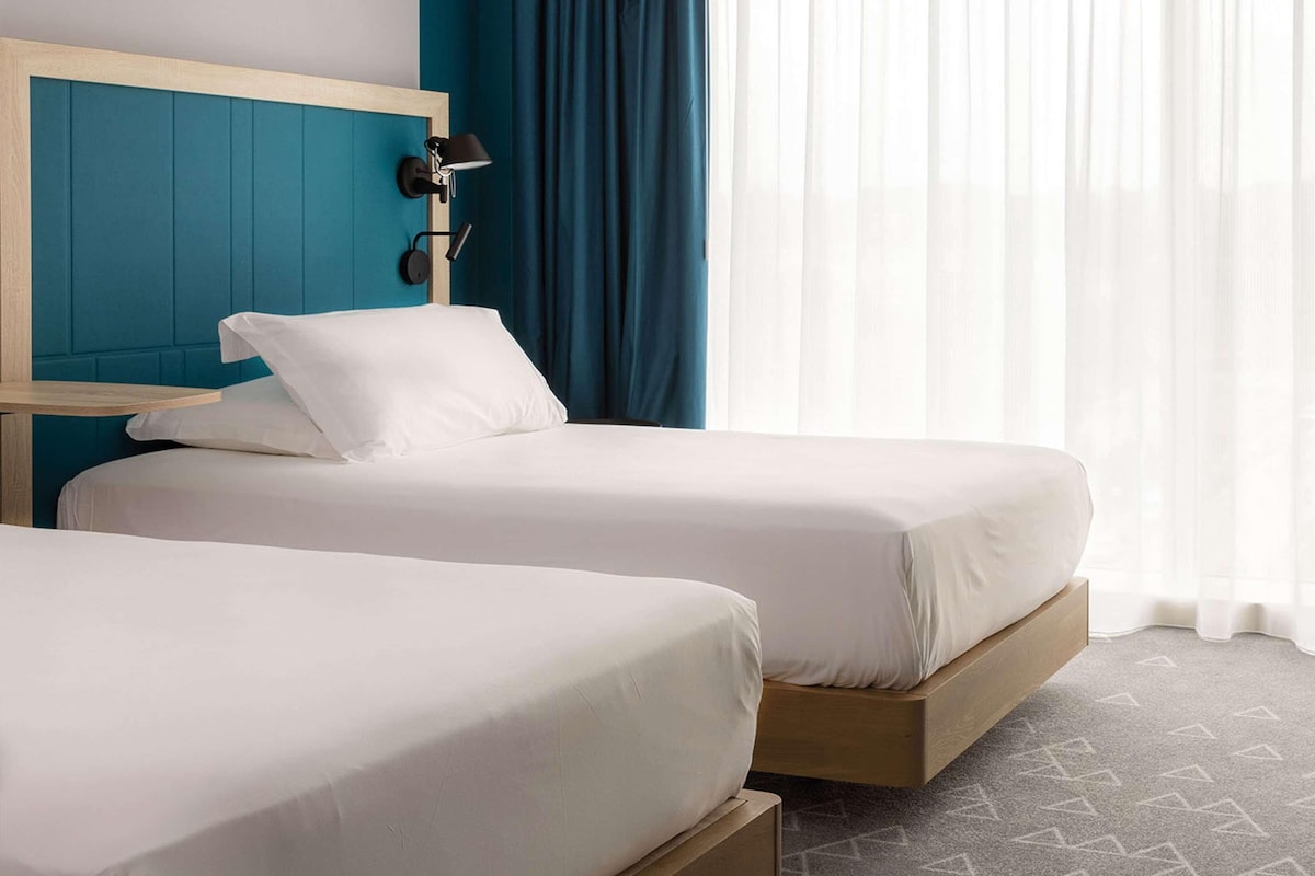 Habitación de hotel con dos camas individuales, cabecera de madera, mesita de noche, lámpara y ventana grande con cortinas traslúcidas.