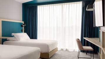 A hotel room with two beds, a desk, a chair, a TV, and a large window with curtains.