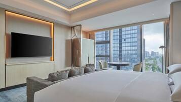 Una habitación de hotel moderna con una cama grande, un televisor de pantalla plana montado en la pared, una zona de estar con sofás y vistas a la ciudad a través de amplias ventanas.