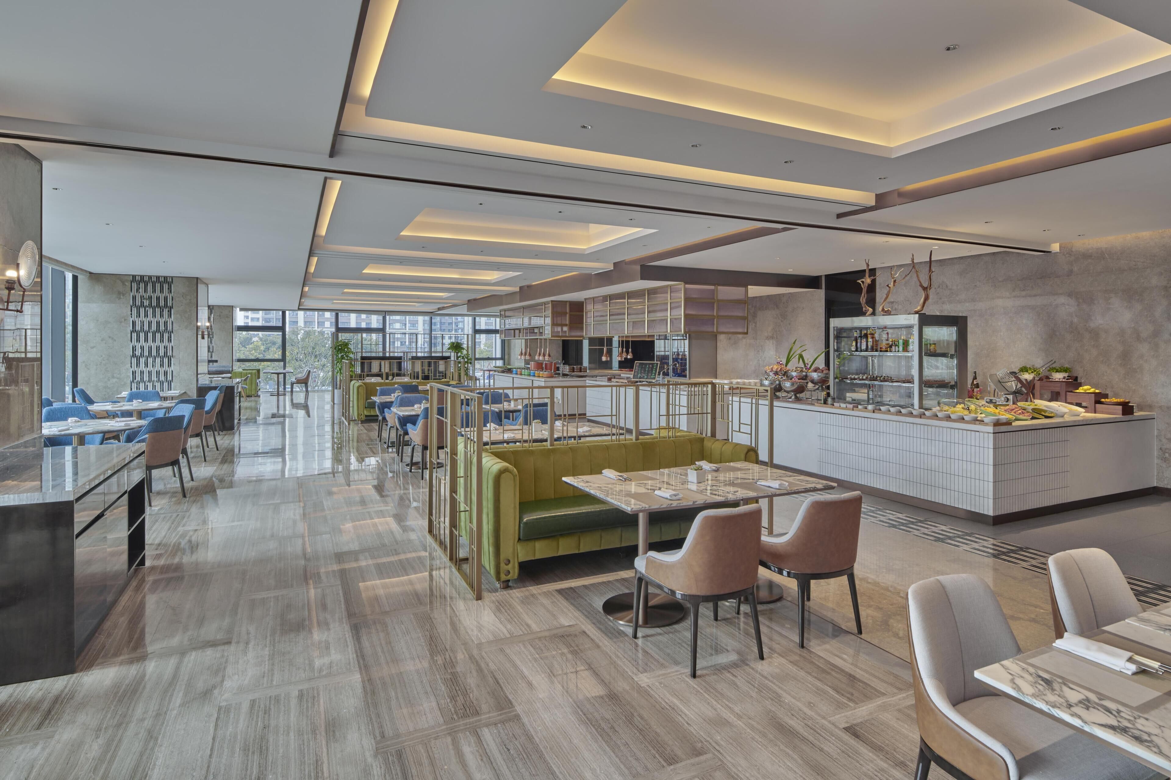 Um hall de hotel moderno com área de estar central, um bar e uma cozinha visíveis ao fundo.