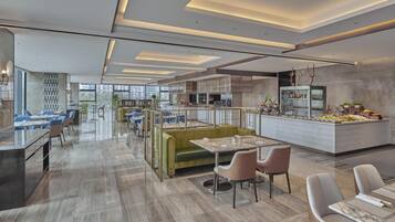 Um hall de hotel moderno com área de estar central, um bar e uma cozinha visíveis ao fundo.