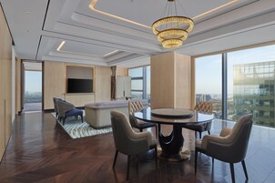 Una habitación de hotel moderna con un amplio ventanal panorámico, una mesa de comedor redonda con sillas, un sofá y un televisor.