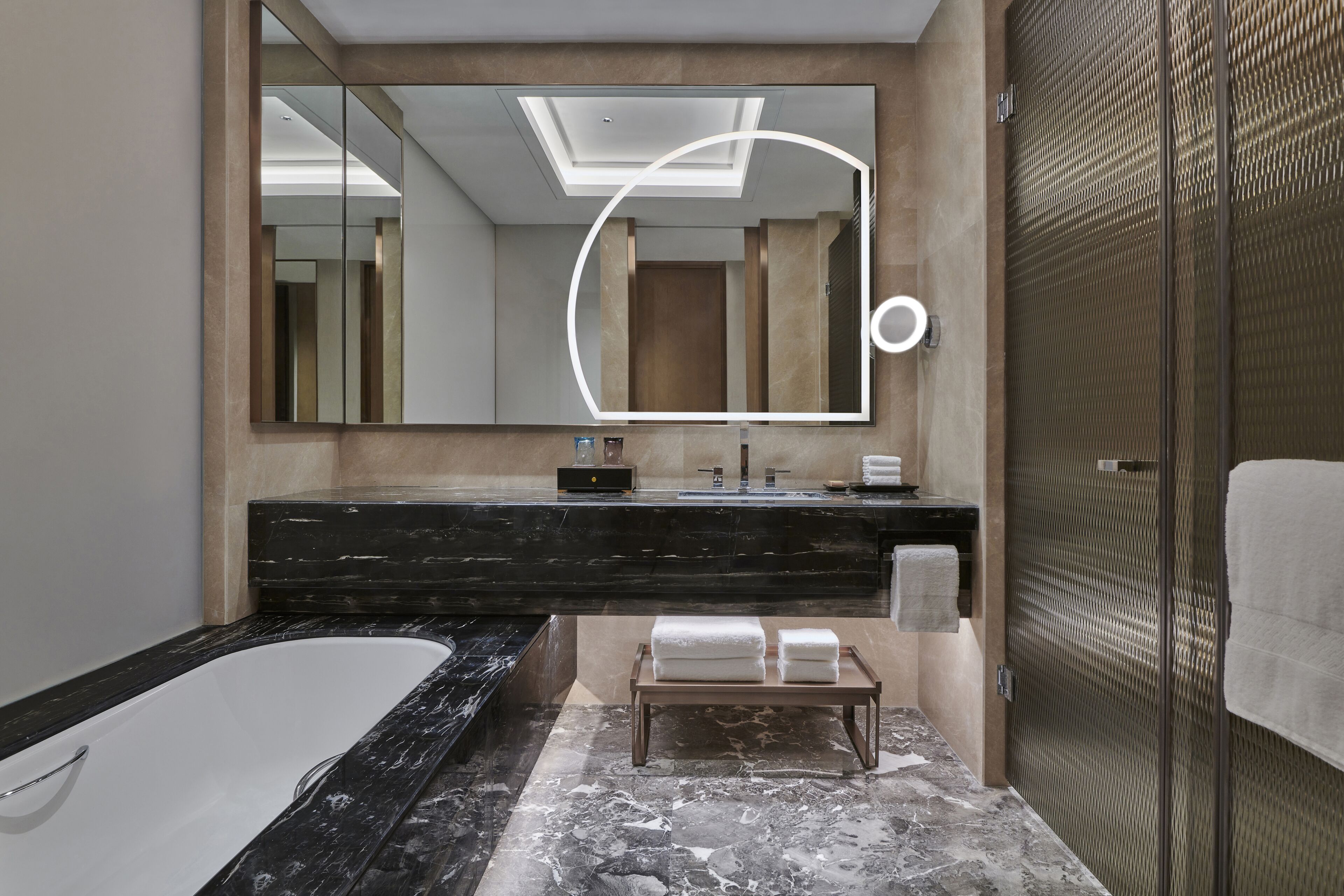Un baño moderno con un espejo grande, una bañera, un tocador con encimera de mármol negro y un banco de madera con toallas.
