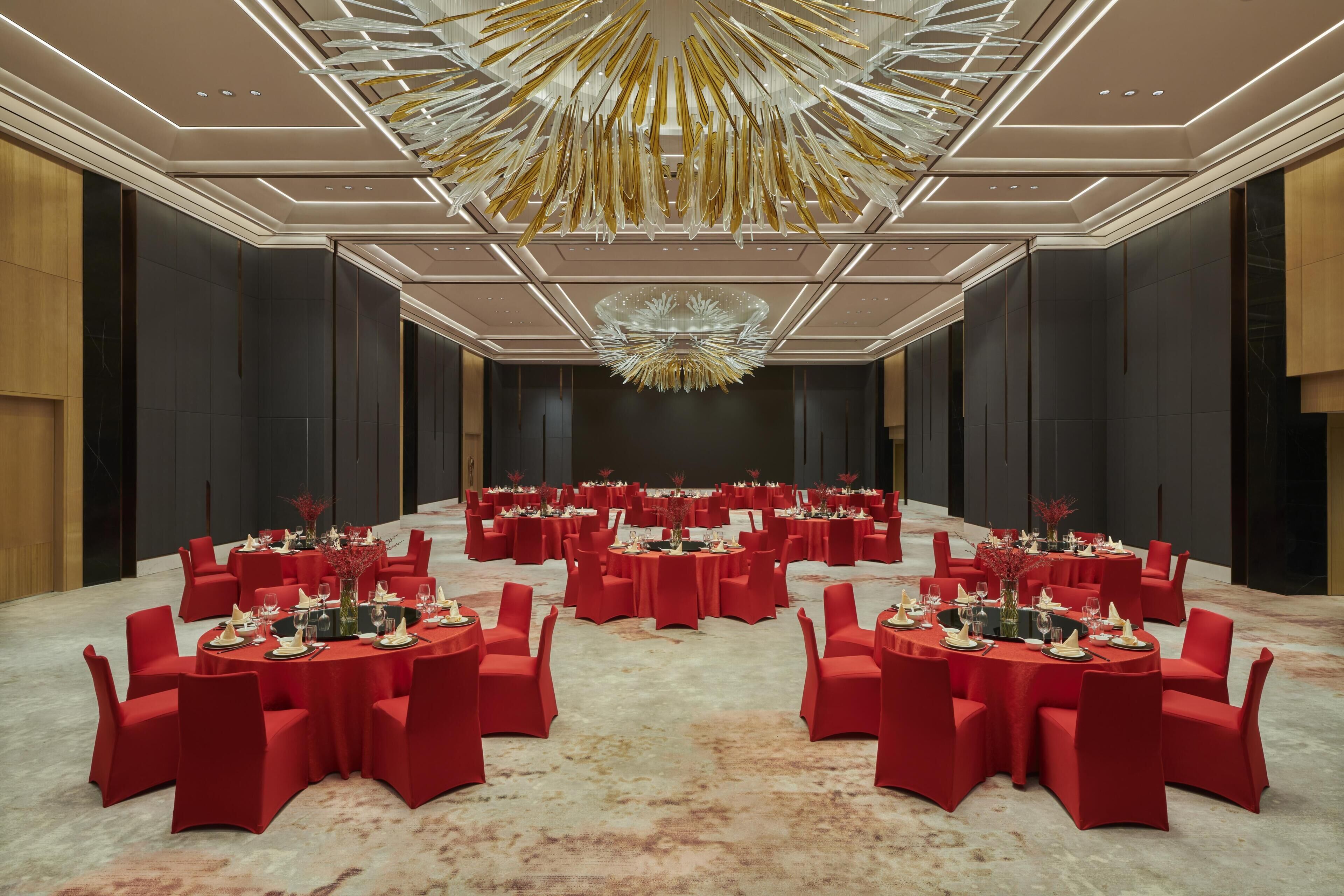A large conference room with multiple round tables, red chairs, and a modern chandelier.