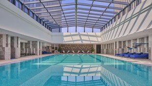 A large indoor swimming pool with a glass ceiling, marble walls, and blue lounge chairs. - Hilton Yancheng (Yancheng)