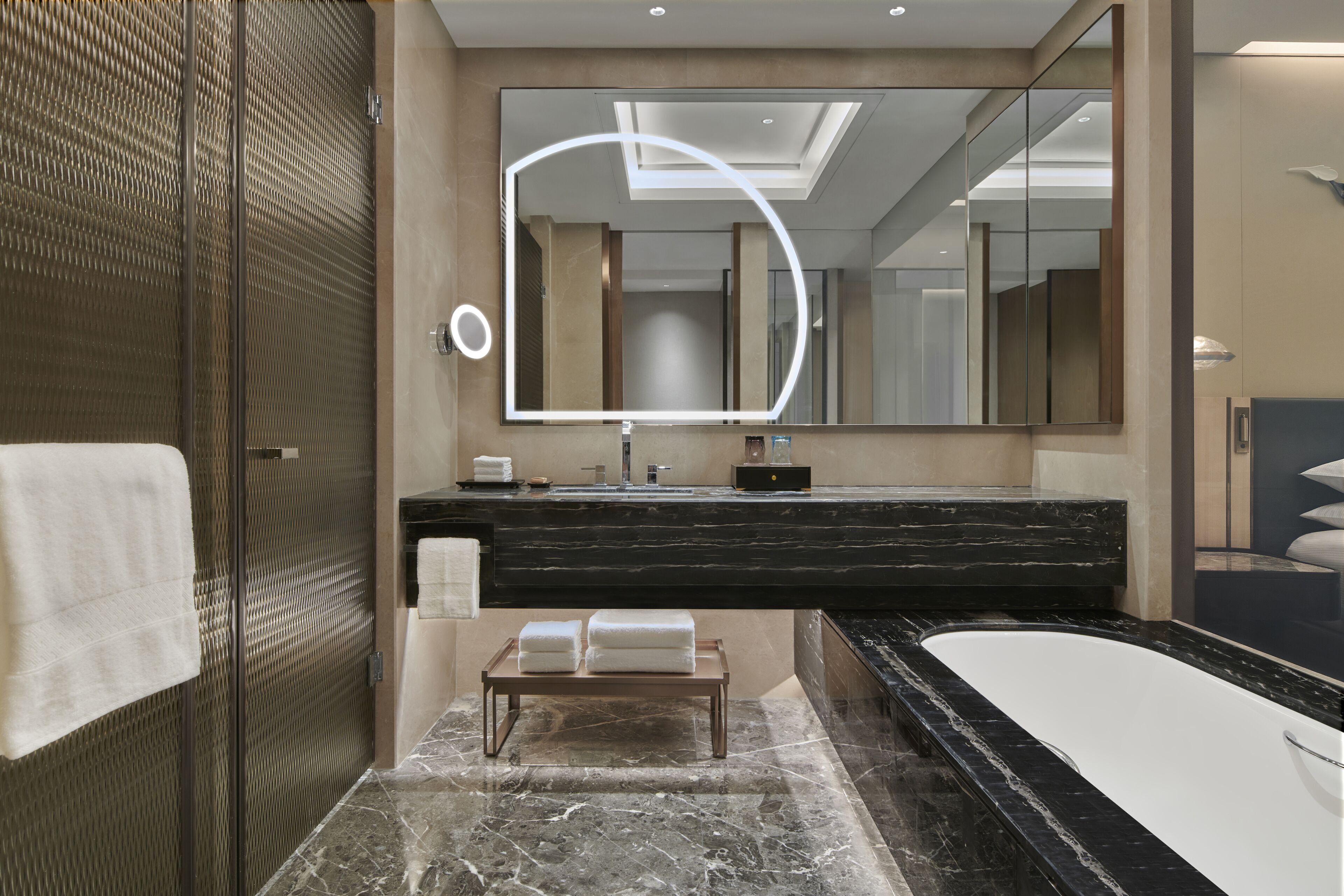 A modern bathroom with a large mirror, a black marble countertop, a bathtub, and a bench with towels.
