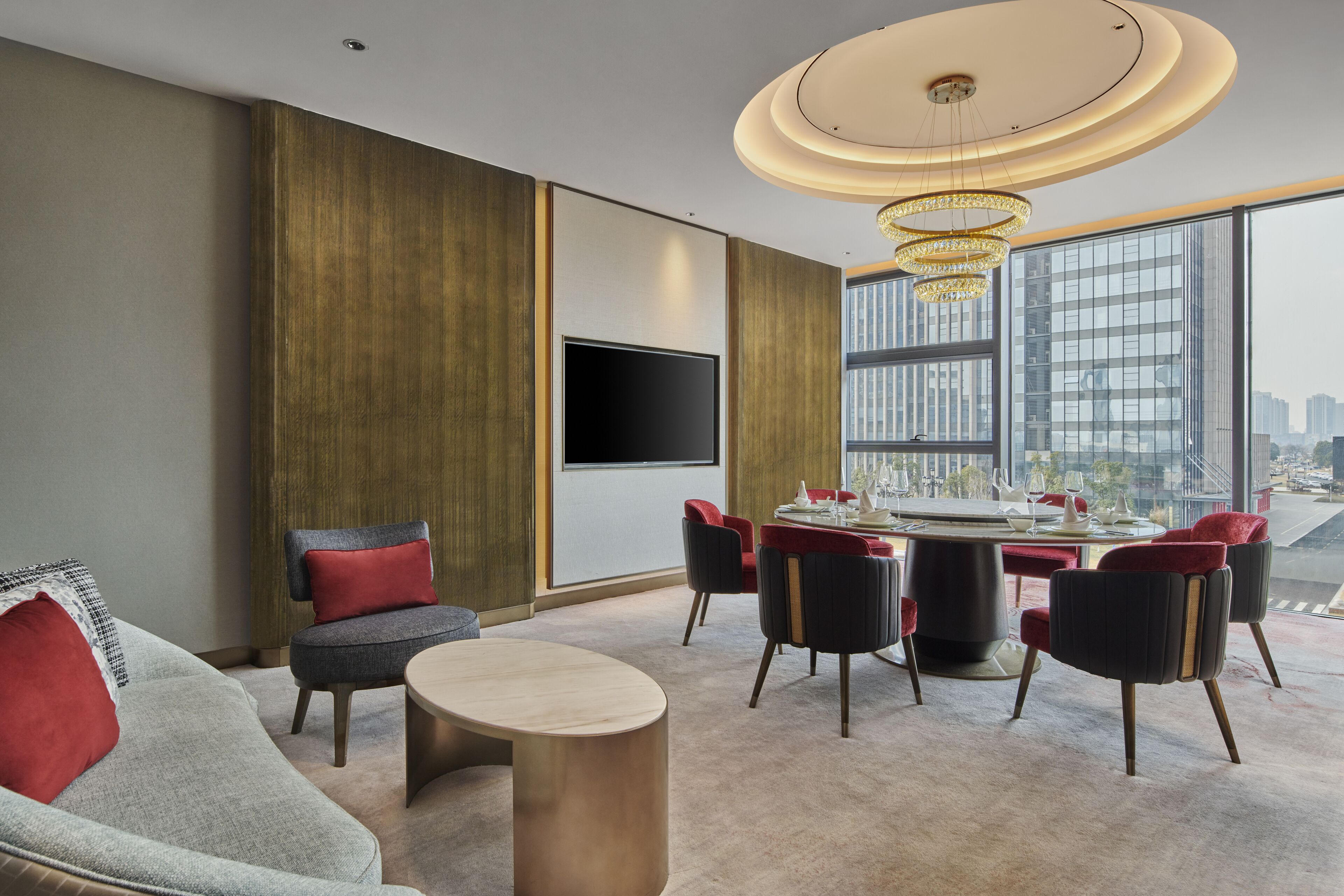 A modern hotel room with a large window, a flat-screen TV, a dining area with red upholstered chairs, and a round coffee table.