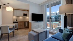 Quarto de hotel moderno com cozinha, televisão de tela plana, sofá e uma grande janela com vista para a cidade.