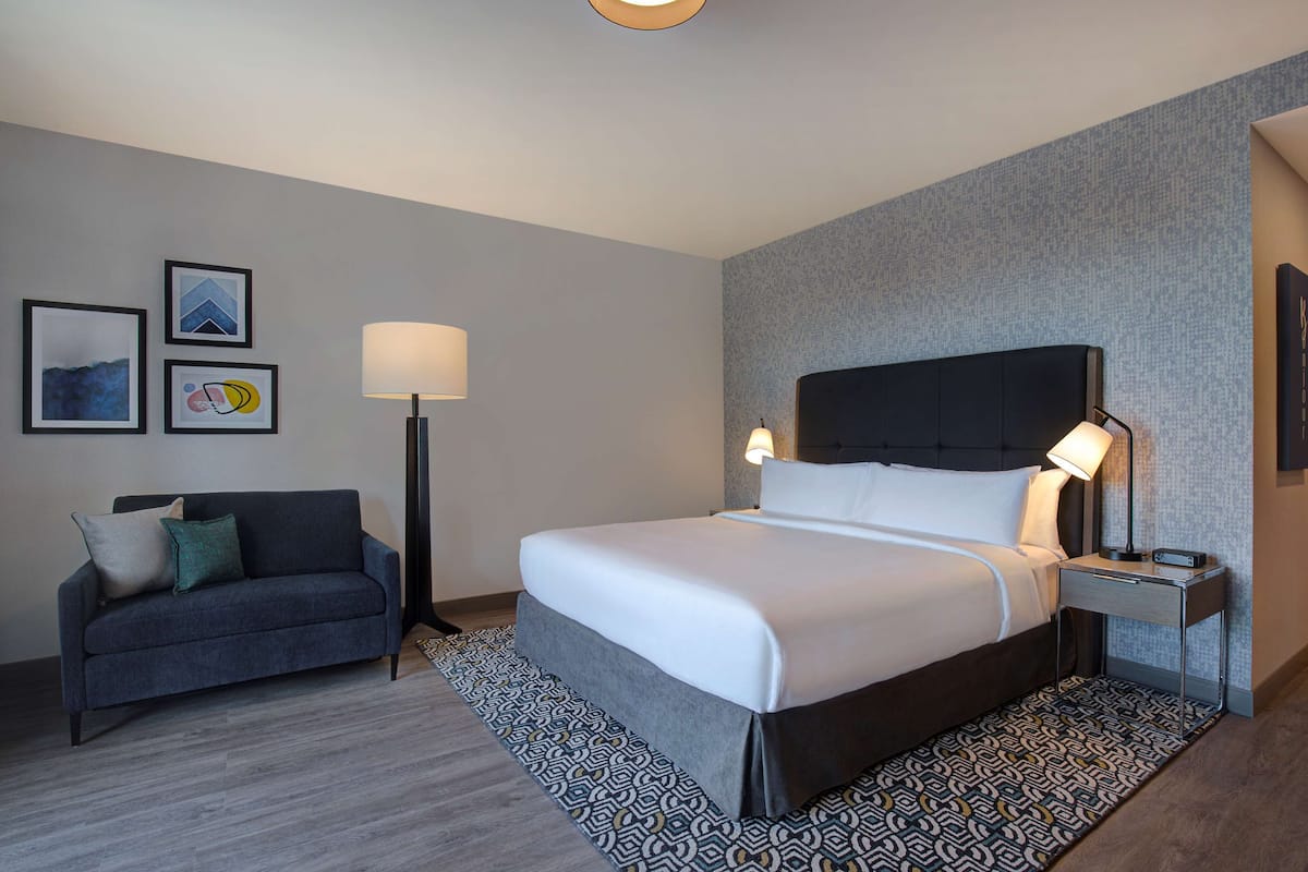 A hotel room with a bed, a desk, a chair, a lamp, and a patterned rug.
