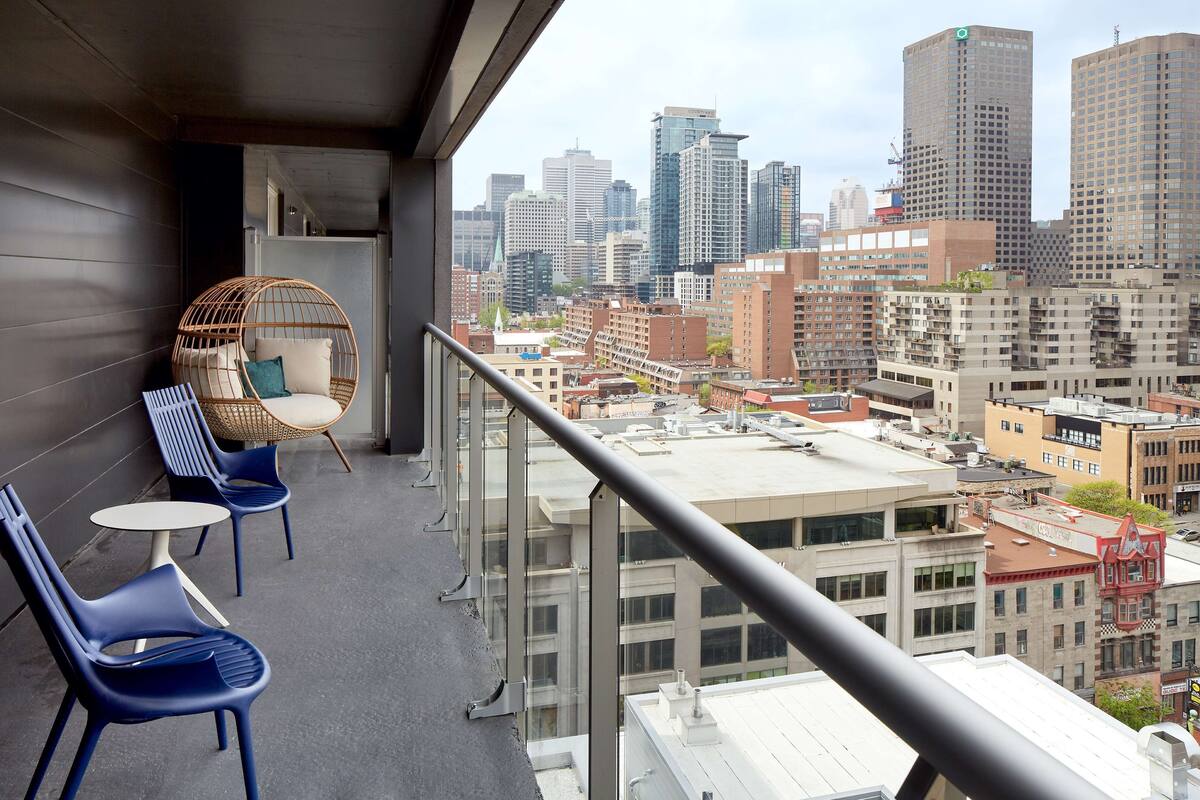 A balcony with wicker chairs and a small table, overlooking a cityscape with various buildings.