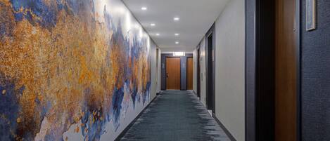 A long, narrow hallway with a textured wall on one side and a plain wall on the other. There are doors on both sides, and the hallway leads to a room at the end.