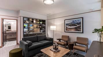 A modern hotel room with a grey sofa, a wooden coffee table, a dining table with chairs, a kitchen area, and a framed picture on the wall.