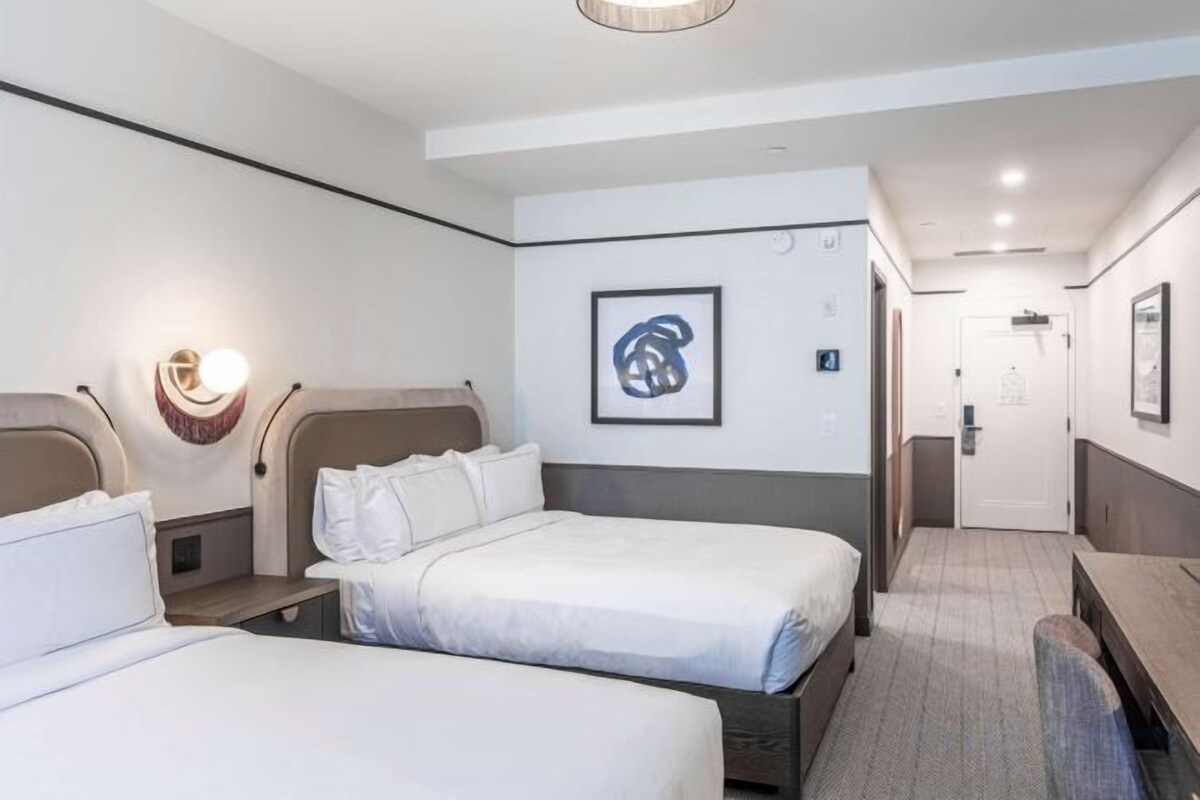 Ein Hotelzimmer mit zwei Betten, einem Schreibtisch und einem Stuhl. Ein gerahmtes Kunstwerk befindet sich an der Wand, und eine Tür führt in einen weiteren Raum.