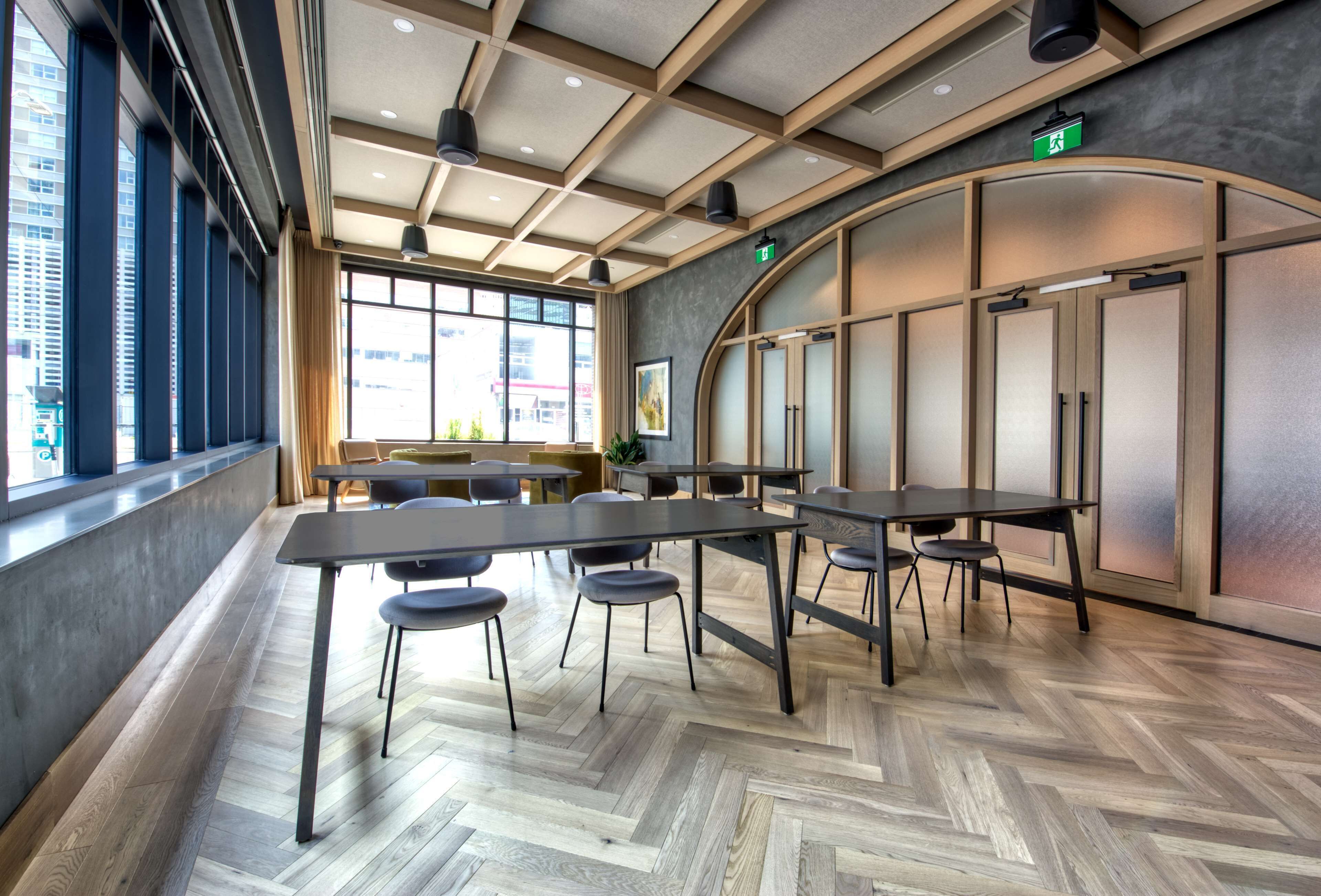 Una sala de conferencias moderna con mesas y sillas grandes, suelo de madera en diseño espiga y grandes ventanales con vistas a la ciudad.