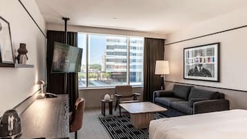 Una habitación de hotel con cama, sofá, escritorio con lámpara, televisor y vistas a la ciudad.