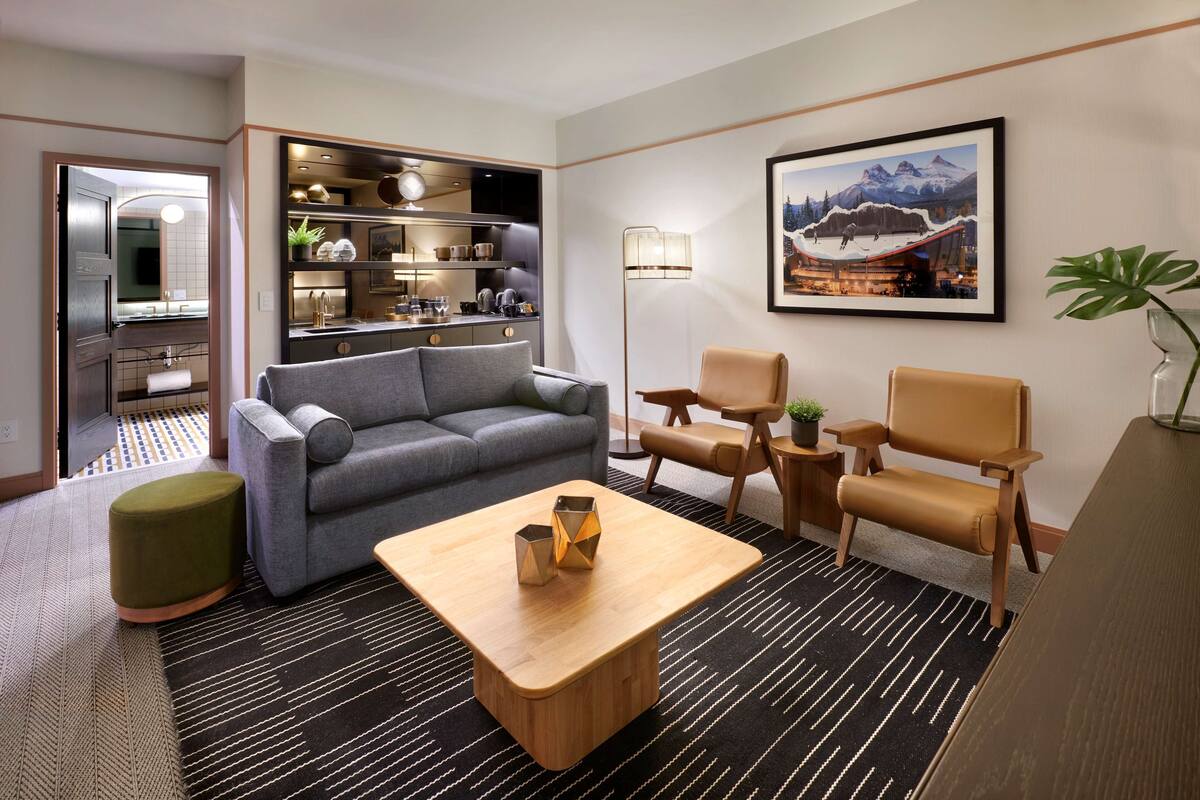 A modern hotel room with a grey sofa, a wooden coffee table, and a dining area with a table and chairs.