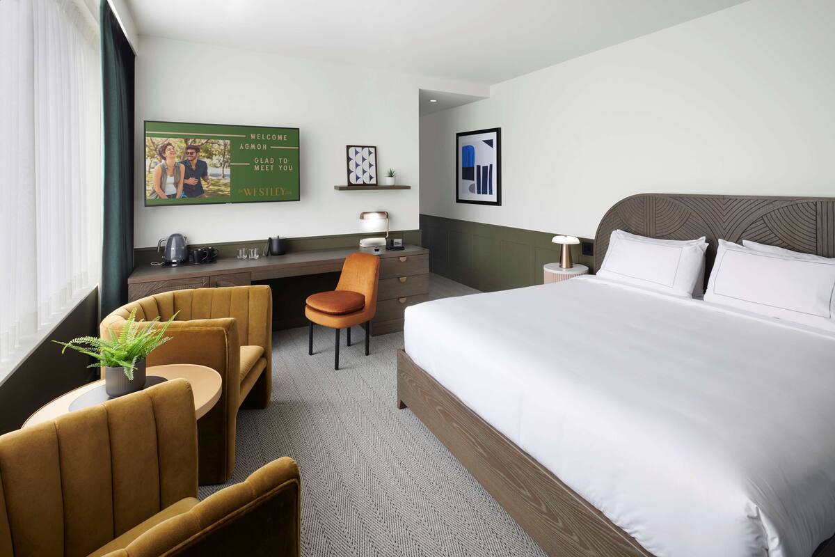 A hotel room with a large bed, a desk with a chair, a small table with a plant, and a framed welcome sign on the wall.