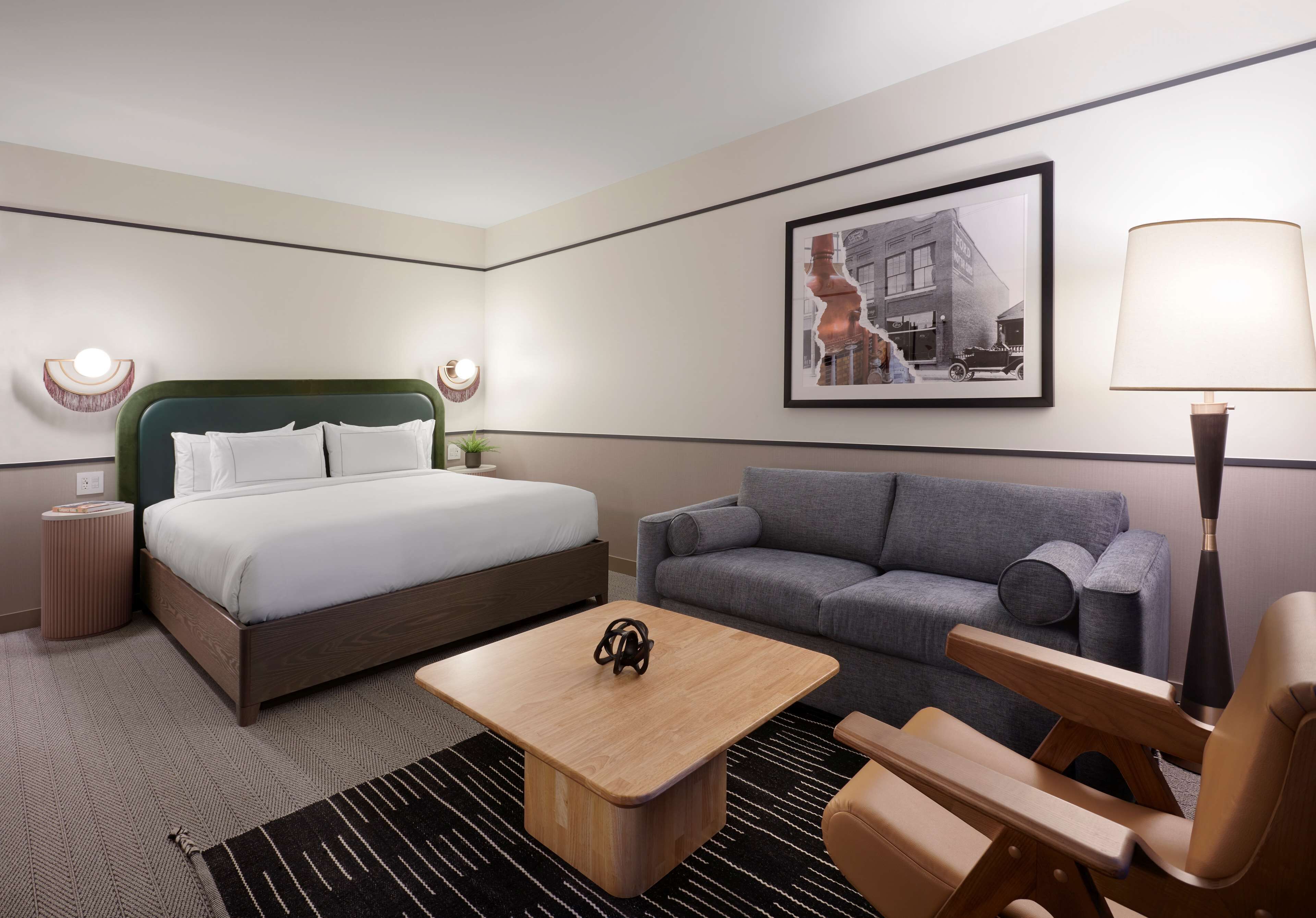 A hotel room with a bed, a gray sofa, a wooden coffee table, a lamp, and a framed picture on the wall.