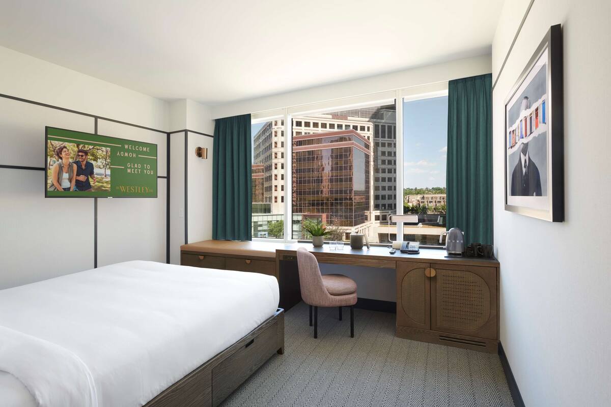 A hotel room with a bed, desk, chair, and large window with curtains.