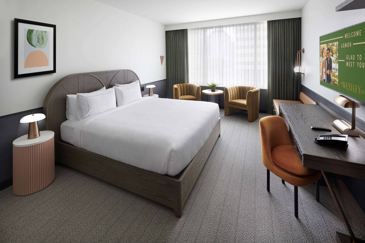 A hotel room with a large bed, two armchairs, a small round table, a desk with a chair, a lamp, and a framed welcome sign.