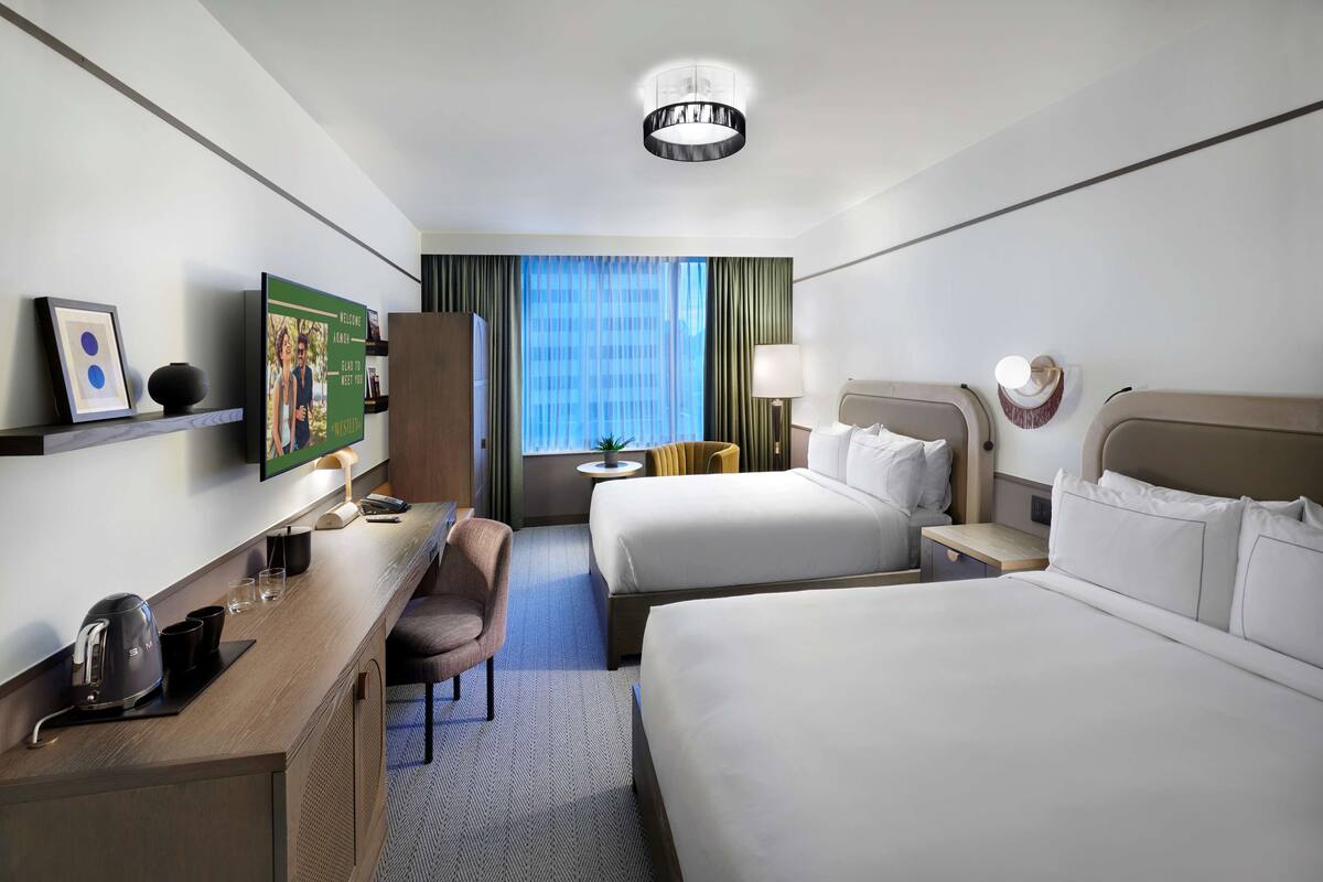 A hotel room with two beds, a desk with a coffee maker, a chair, a TV, and a window with curtains.