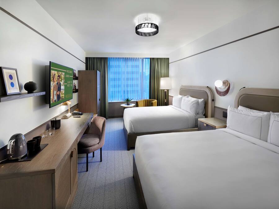 Habitación de hotel con dos camas, un escritorio con cafetera, una silla, televisor y ventana con cortinas.