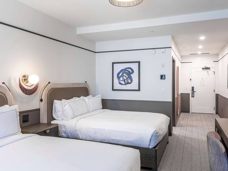 A hotel room with two beds, a desk, and a chair. There is a framed artwork on the wall and a door in the background.