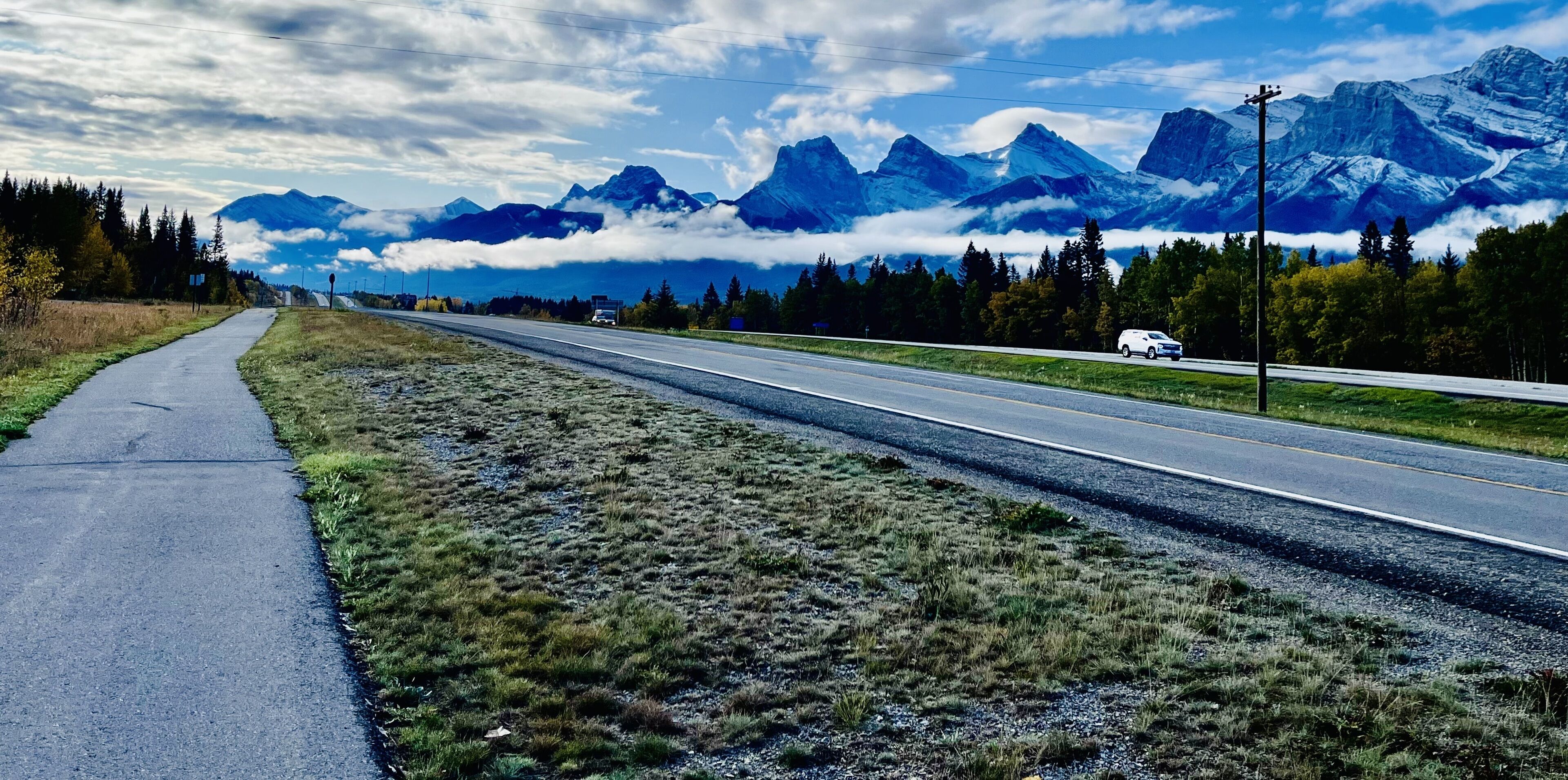 Une route bordée d’un trottoir sur le côté gauche, entourée d’herbe et d’arbres, qui mène à une chaîne de montagnes.