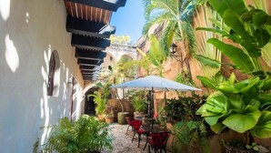 An outdoor patio area with stone flooring, red and black chairs, a white umbrella, and lush tropical plants. - El Beaterio (Santo Domingo)