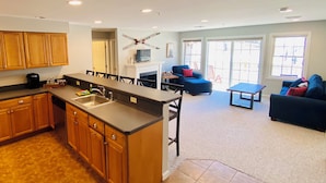 A modern kitchen with wooden cabinets and a large island, a dining area with a blue chair and a small table, and a living room with a blue sofa and a fireplace.