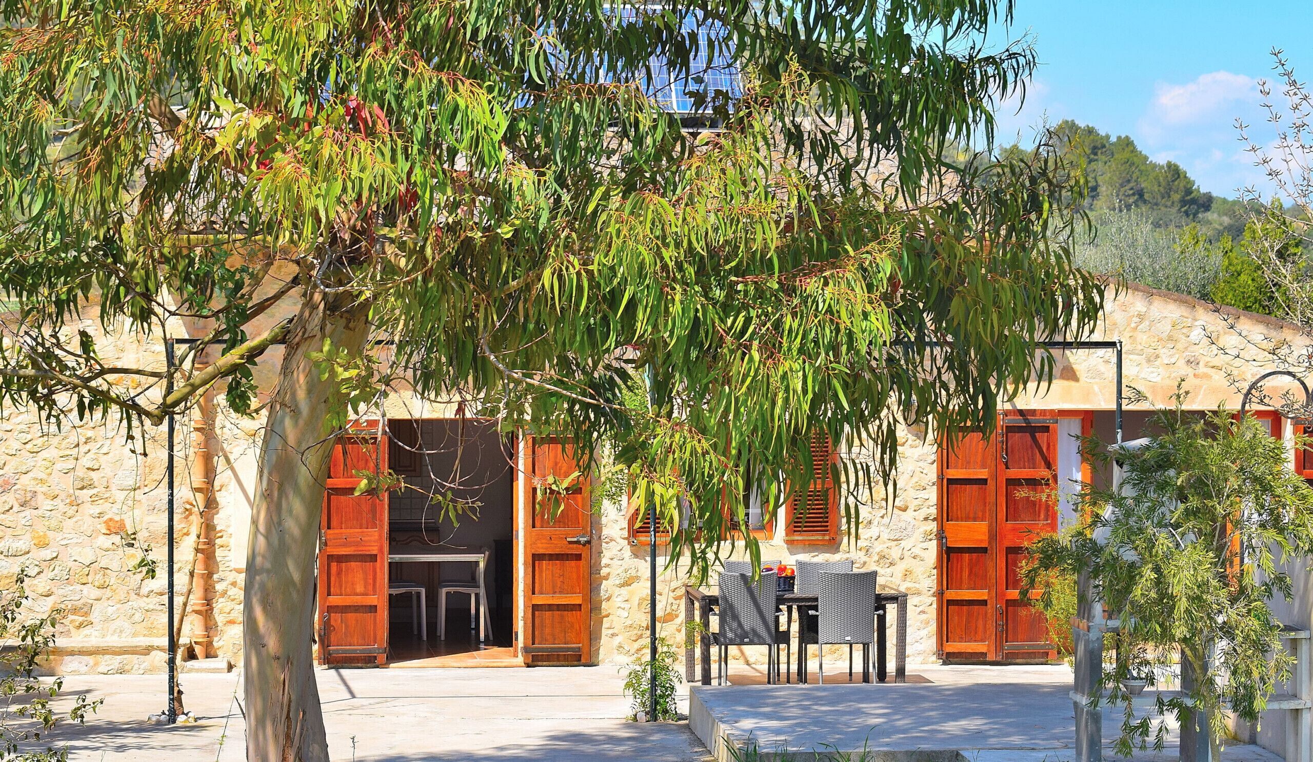A stone building with wooden shutters, a stone staircase, and a table with chairs outside.