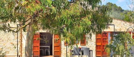 A stone building with wooden shutters, a stone staircase, and a table with chairs outside.