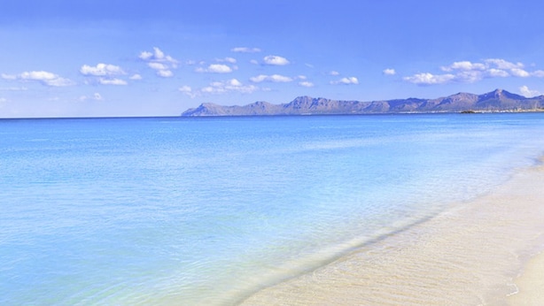 Ein Strand mit klarem, blauem Wasser und einem Bergmassiv im Hintergrund.