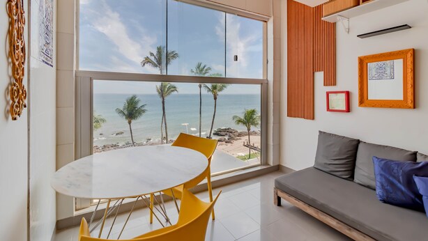 A modern room with a large window overlooking a beach, a round table with yellow chairs, and a grey sofa with blue pillows. - RV03 Quarto e Sala Vista Mar (Salvador)